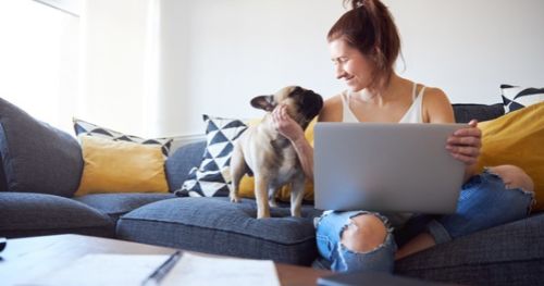 A woman on her laptop, smiling. Her dog sits beside her.